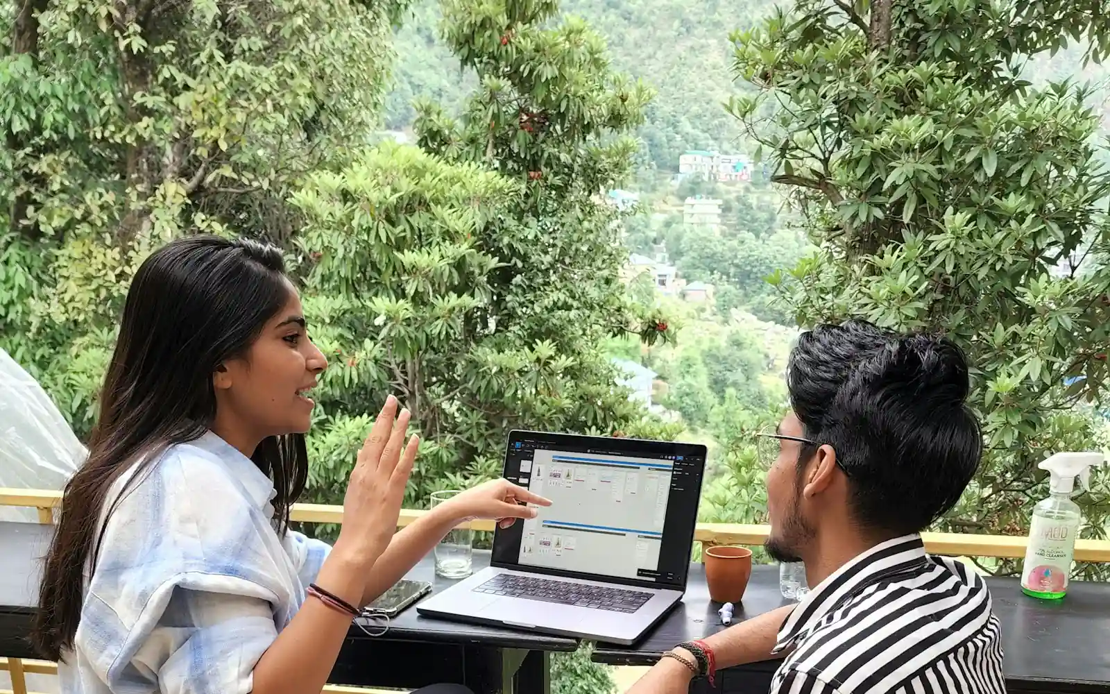 A woman and a man discuss a dashboard on a laptop at an outdoor cafe table with forested hills behind them in Himachal Pradesh, India