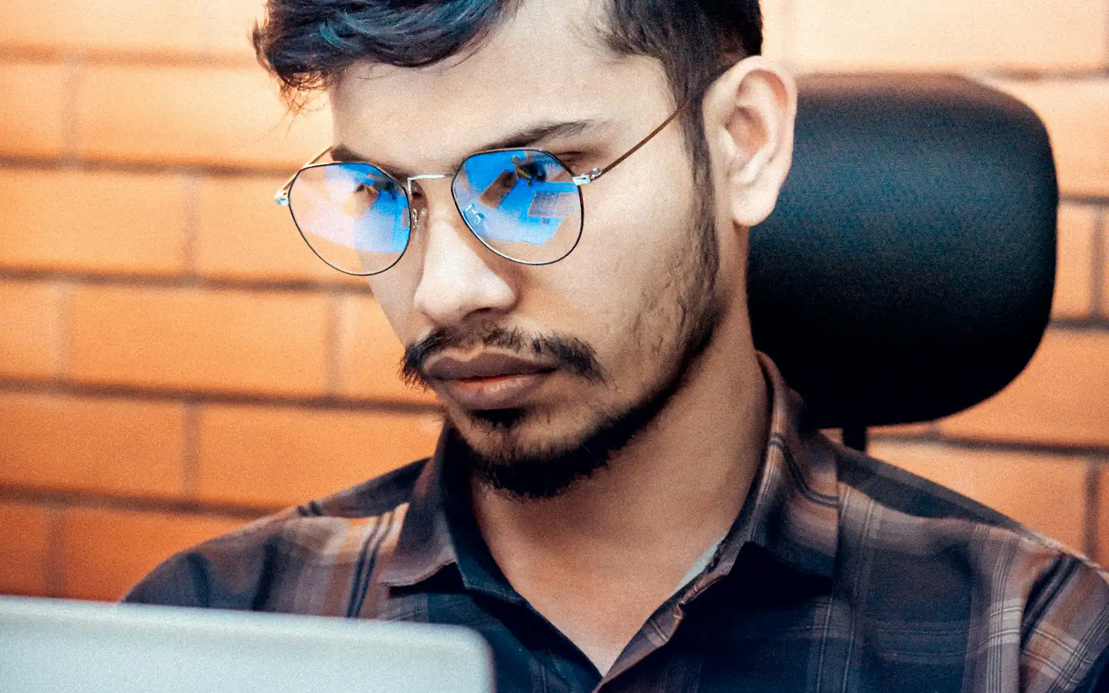 A young South Asian man in glasses and a checked shirt studies his laptop screen at an office chair against a brick wall