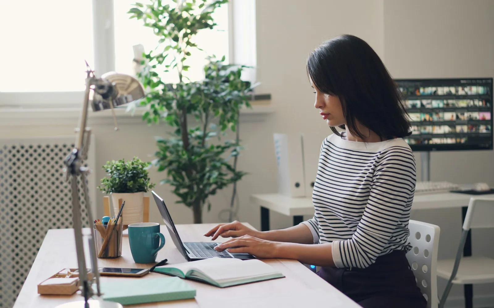Woman typing on a laptop at a bright office desk with a notebook, coffee mug and plants, reviewing work on screen