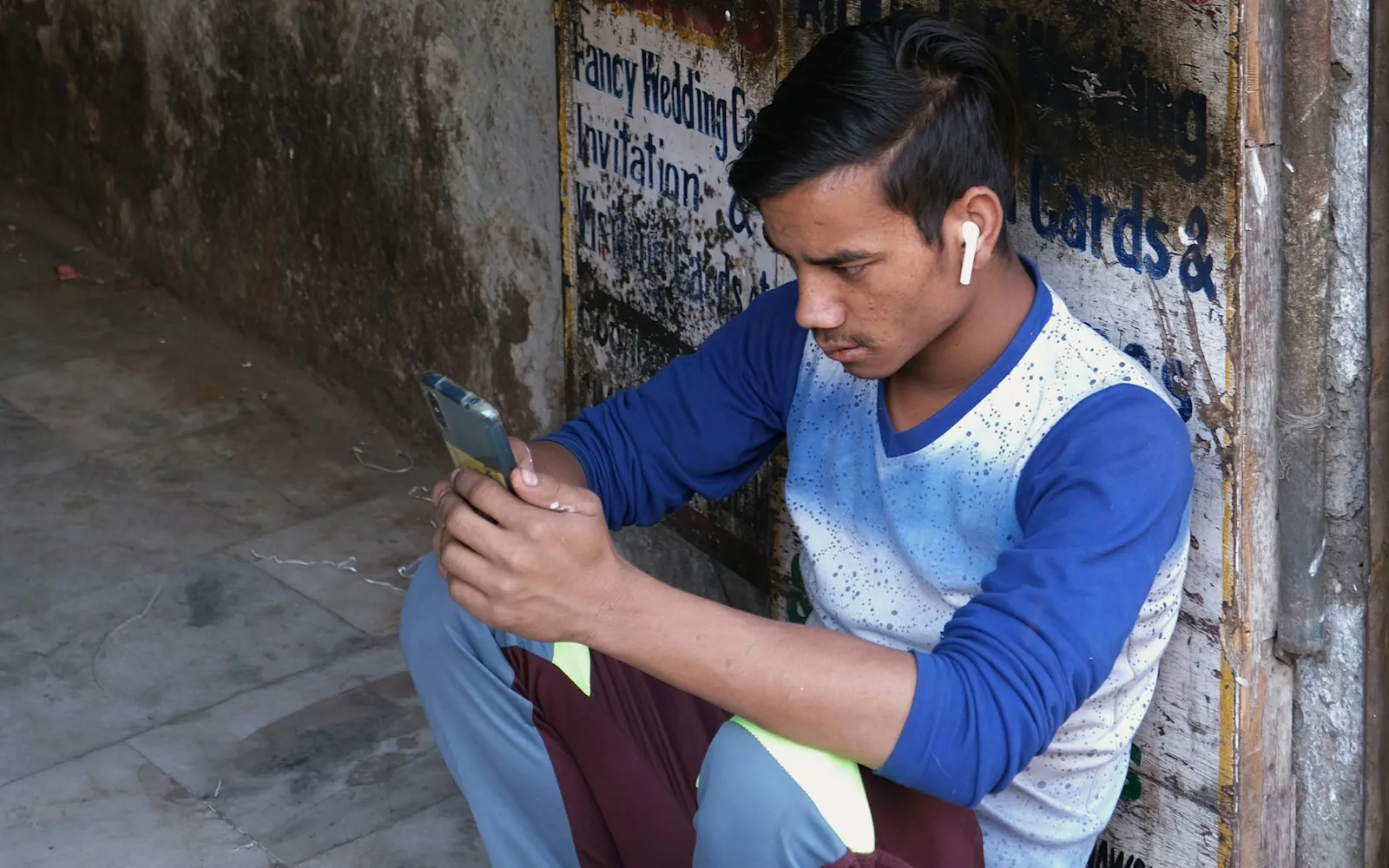 Young man in a blue and white shirt sitting against painted shop signs in Old Delhi, looking closely at his smartphone