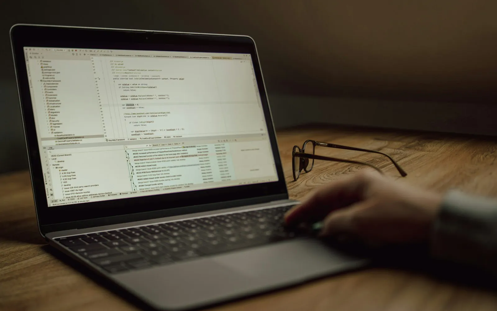Hand on a laptop trackpad with lines of code on screen and reading glasses resting on a wooden desk in low light
