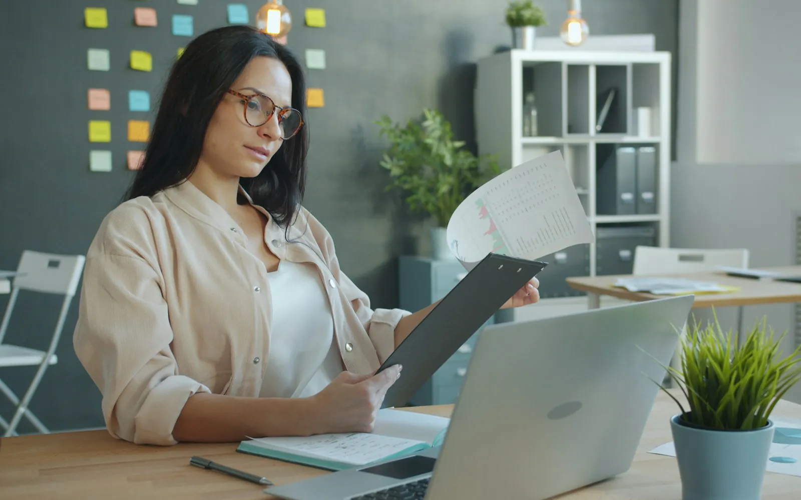 Woman in glasses reviewing a printed chart on a clipboard beside an open laptop at an office desk with sticky notes on the wall