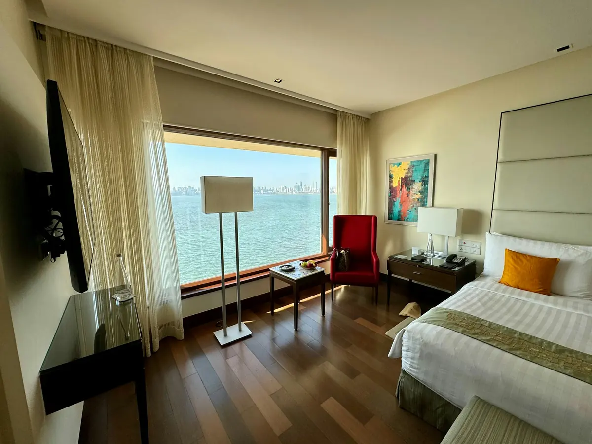Hotel room in Mumbai with a large window overlooking the Arabian Sea and city skyline, bed, armchair and floor lamp