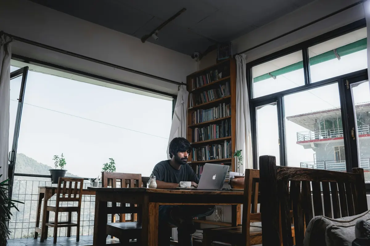 Man working on a laptop at a wooden table in a bright cafe with bookshelves and mountain views in Dharamshala